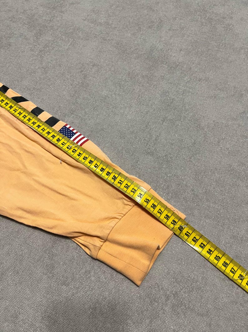 May include: A long-sleeved, peach-colored shirt sleeve with a black and white stripe design and a small American flag patch. A yellow measuring tape is laid across the sleeve, showing measurements in inches.