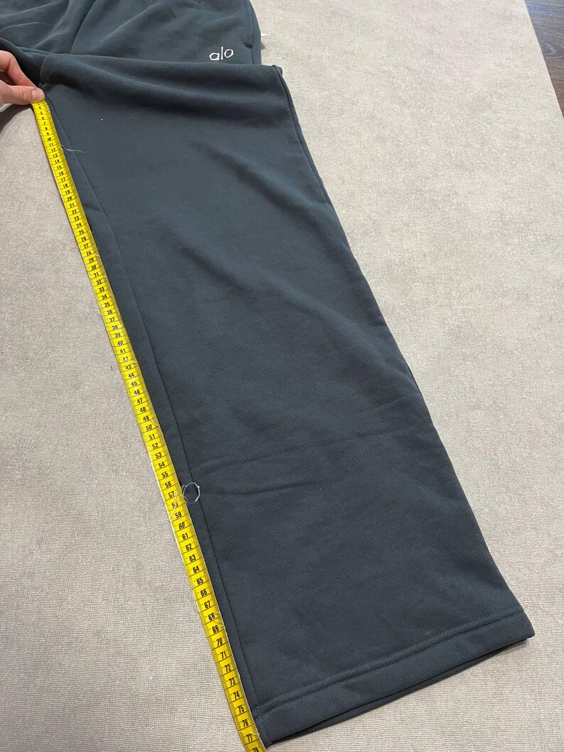 May include: Dark charcoal gray sweatpants featuring the white text 'alo' near the waistband. A yellow measuring tape is placed along the side seam, showing a length measurement extending past 30 inches on the tape's imperial scale.