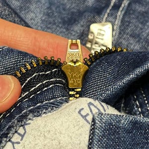 Puede incluir: Primer plano de una cremallera dorada de una prenda vaquera. El tirador de la cremallera est&aacute; marcado con "YKK 4YG". La tela vaquera es de un lavado azul medio con costuras blancas. Una etiqueta de tela con la palabra "FATIO" es visible.