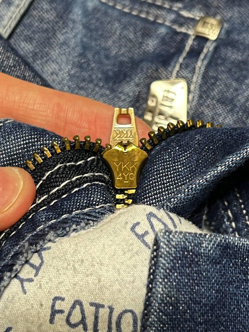Puede incluir: Primer plano de una cremallera dorada de una prenda vaquera. El tirador de la cremallera est&aacute; marcado con "YKK 4YG". La tela vaquera es de un lavado azul medio con costuras blancas. Una etiqueta de tela con la palabra "FATIO" es visible.