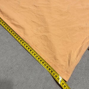 May include: A peach-colored fabric is measured with a yellow measuring tape. The tape shows measurements up to 62 inches. The fabric has visible creases and folds, suggesting it is a piece of clothing or textile.