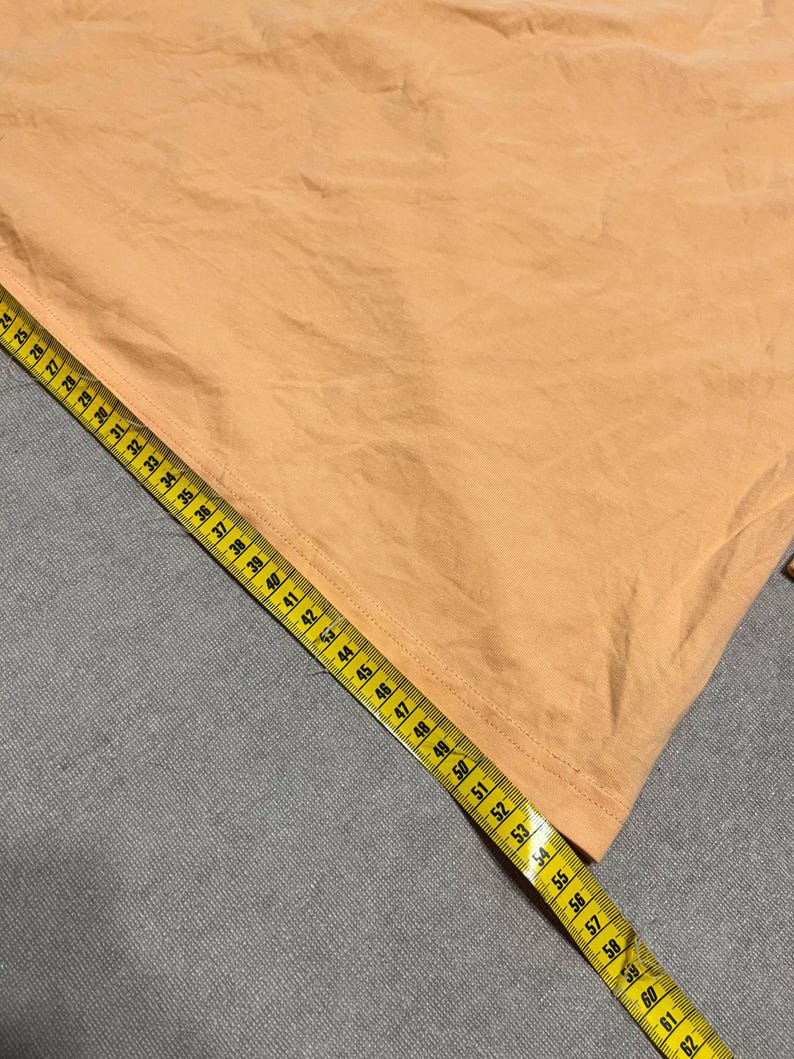 May include: A peach-colored fabric is measured with a yellow measuring tape. The tape shows measurements up to 62 inches. The fabric has visible creases and folds, suggesting it is a piece of clothing or textile.