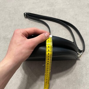 May include: Black, round crossbody bag with a long, adjustable strap. The bag is being measured with a yellow measuring tape. The bag's height is approximately 6 inches.