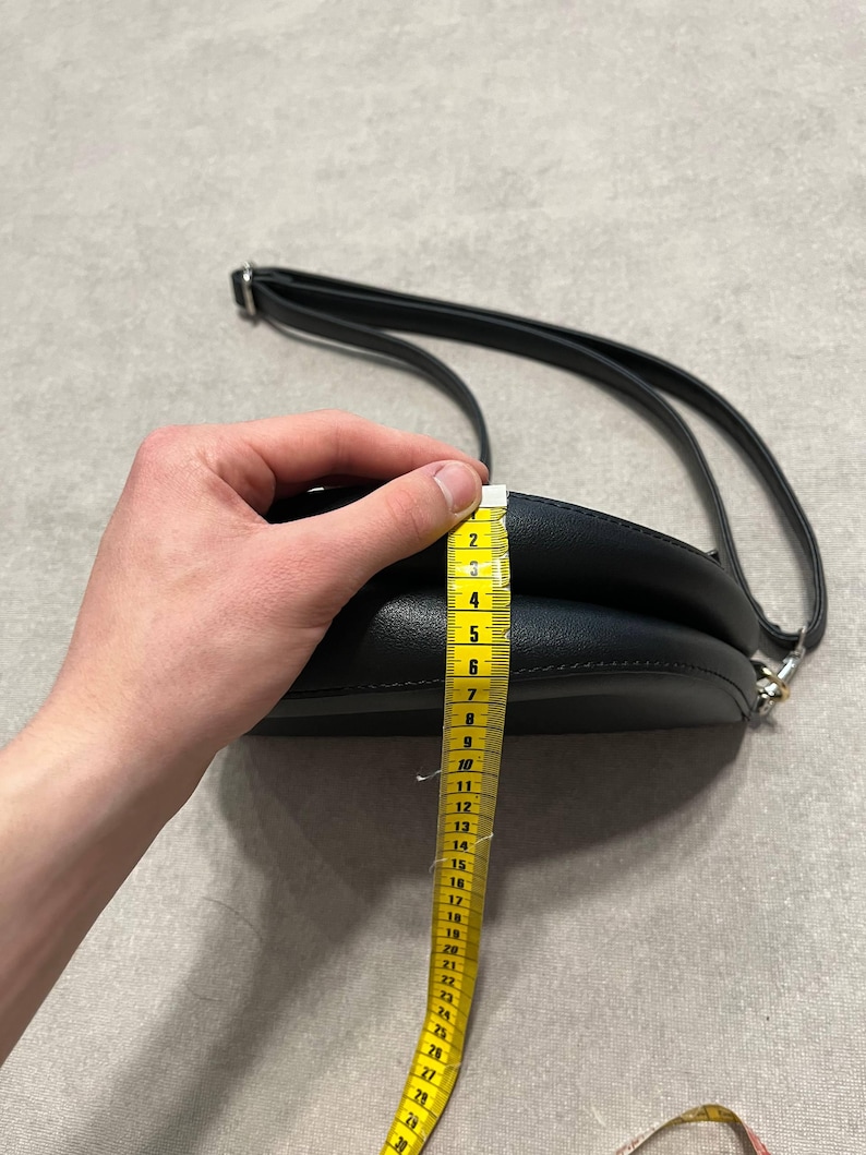 May include: Black, round crossbody bag with a long, adjustable strap. The bag is being measured with a yellow measuring tape. The bag's height is approximately 6 inches.
