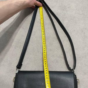 May include: Black crossbody bag with a long strap. The bag is measured with a yellow measuring tape, showing a strap length of approximately 48 inches. The bag has the brand name "VALENTINO" embossed on the front.