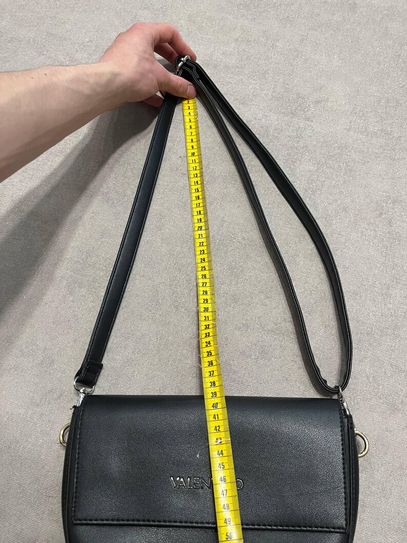May include: Black crossbody bag with a long strap. The bag is measured with a yellow measuring tape, showing a strap length of approximately 48 inches. The bag has the brand name "VALENTINO" embossed on the front.