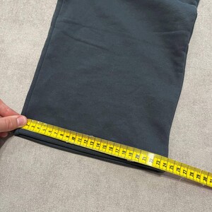 May include: A pair of dark gray sweatpants with a straight leg opening. A yellow measuring tape is placed across the hem, showing a width of approximately 9.5 inches from the edge to the seam, indicating the flat measurement of the leg opening.