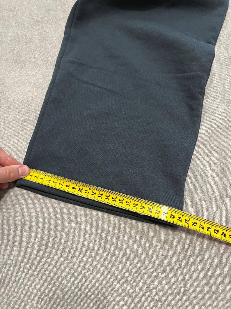 May include: A pair of dark gray sweatpants with a straight leg opening. A yellow measuring tape is placed across the hem, showing a width of approximately 9.5 inches from the edge to the seam, indicating the flat measurement of the leg opening.