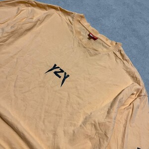 May include: A light orange long-sleeve shirt with the letters "YZY" in black on the chest. The sleeve has a black graphic. The shirt is made of a soft material.