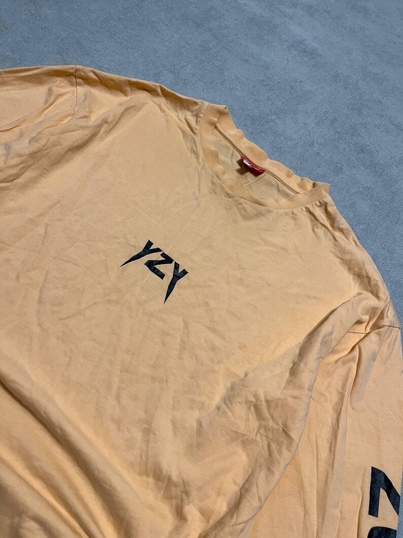 May include: A light orange long-sleeve shirt with the letters "YZY" in black on the chest. The sleeve has a black graphic. The shirt is made of a soft material.