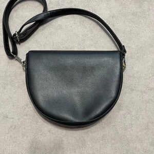 May include: Black crossbody bag with a semi-circular design. The bag features a long, adjustable strap and silver-toned hardware. The bag is made of a smooth, matte material.