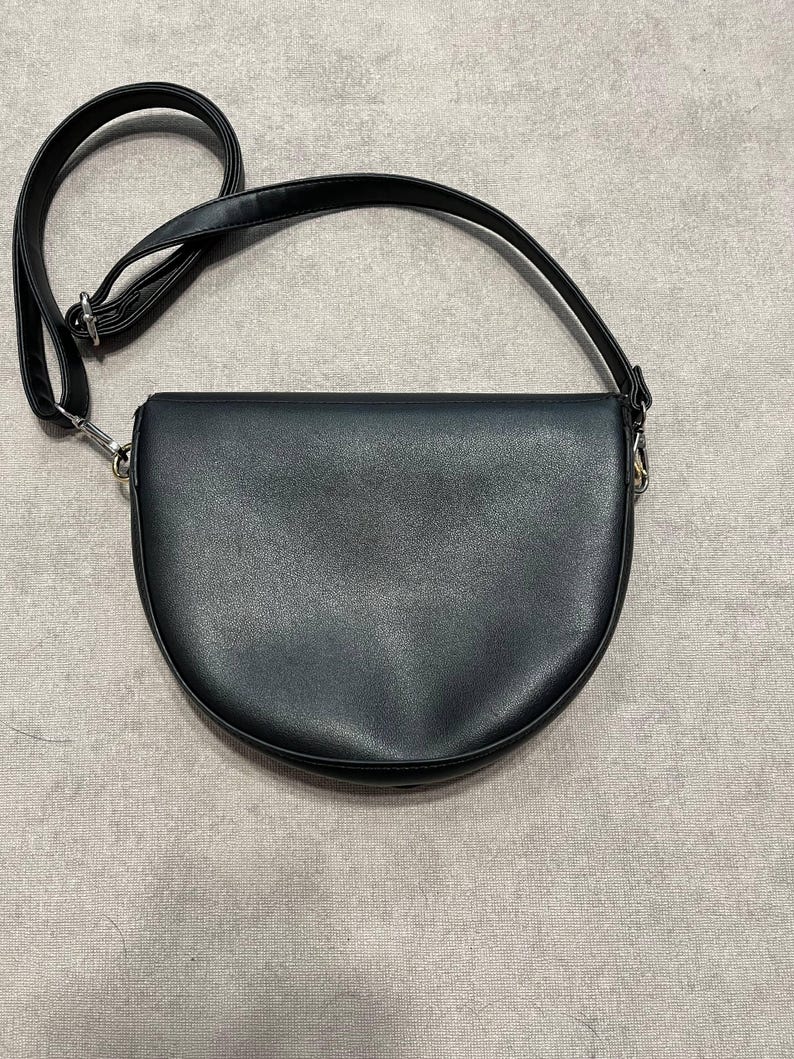 May include: Black crossbody bag with a semi-circular design. The bag features a long, adjustable strap and silver-toned hardware. The bag is made of a smooth, matte material.