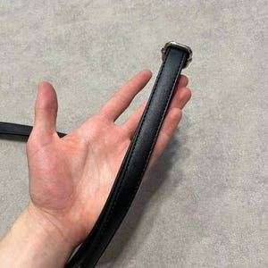 May include: A black leather belt with a silver-toned buckle. The belt is held up by a hand against a gray background. The buckle is square with a decorative design.