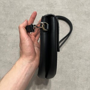 May include: Black crossbody bag with a long strap. The bag is made of a smooth material and features a silver-colored clasp. The strap is thin and appears to be adjustable. The bag is held by a person.