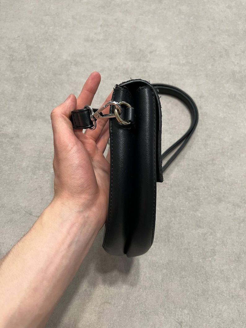 May include: Black crossbody bag with a long strap. The bag is made of a smooth material and features a silver-colored clasp. The strap is thin and appears to be adjustable. The bag is held by a person.