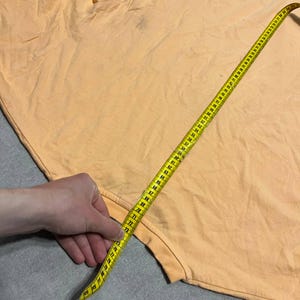 May include: A light orange t-shirt is measured with a yellow measuring tape. The tape is extended across the fabric, showing measurements in inches. The shirt appears to be made of a soft material.
