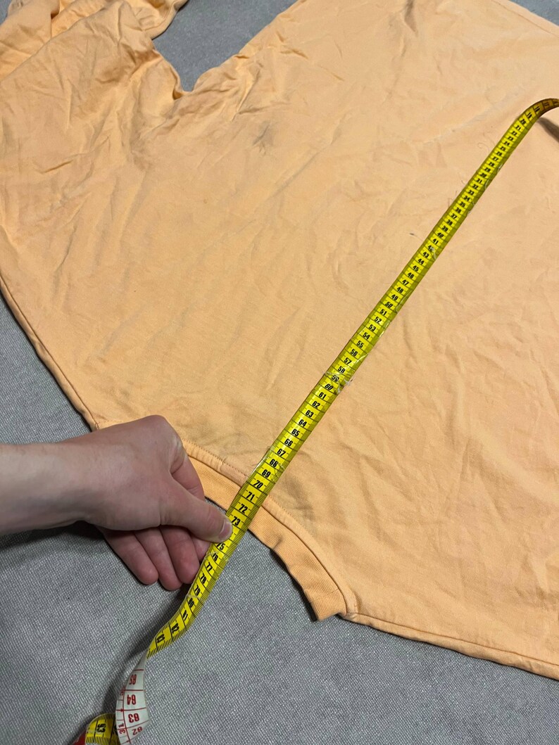 May include: A light orange t-shirt is measured with a yellow measuring tape. The tape is extended across the fabric, showing measurements in inches. The shirt appears to be made of a soft material.