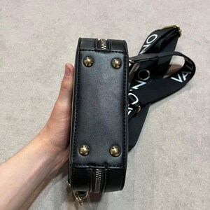 May include: Black rectangular crossbody bag with gold-colored studs and zipper detailing. The bag has a black strap with white text. The bag is held by a person.