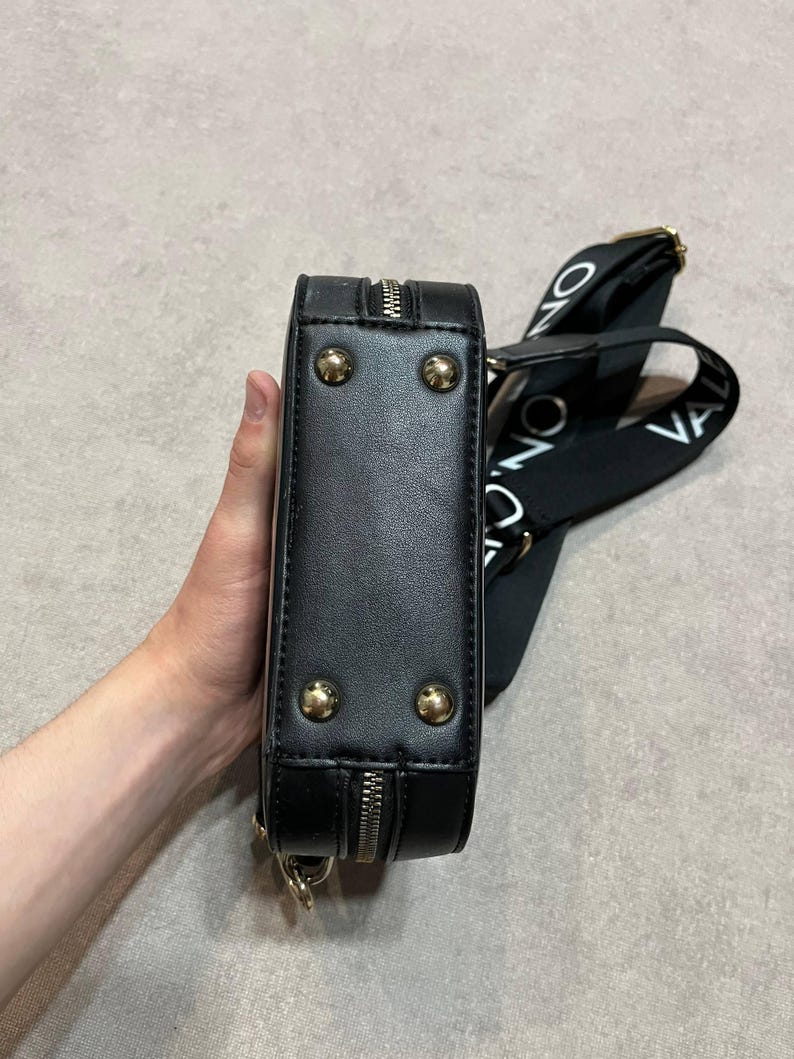 May include: Black rectangular crossbody bag with gold-colored studs and zipper detailing. The bag has a black strap with white text. The bag is held by a person.