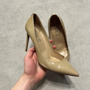 Puede incluir: Un par de zapatos de tac&oacute;n de charol beige con punta. Los zapatos tienen tacones de aguja y la marca es visible dentro del zapato. Los zapatos son sostenidos por una persona.
