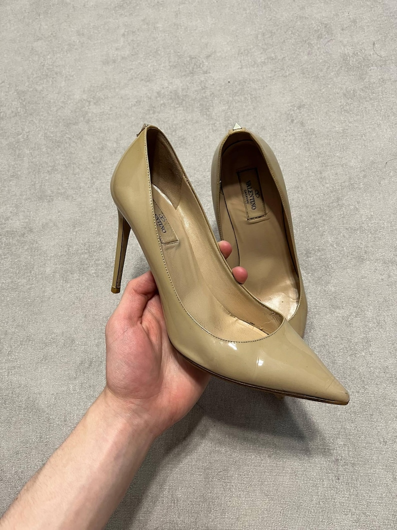 Puede incluir: Un par de zapatos de tac&oacute;n de charol beige con punta. Los zapatos tienen tacones de aguja y la marca es visible dentro del zapato. Los zapatos son sostenidos por una persona.