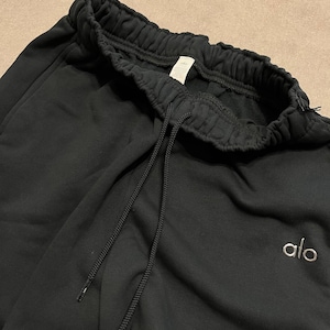 May include: Black sweatpants featuring an elasticized waistband with a matching black drawstring. The garment includes a small white embroidered logo reading alo on the left side. The fabric has a soft, matte texture and a relaxed fit design.
