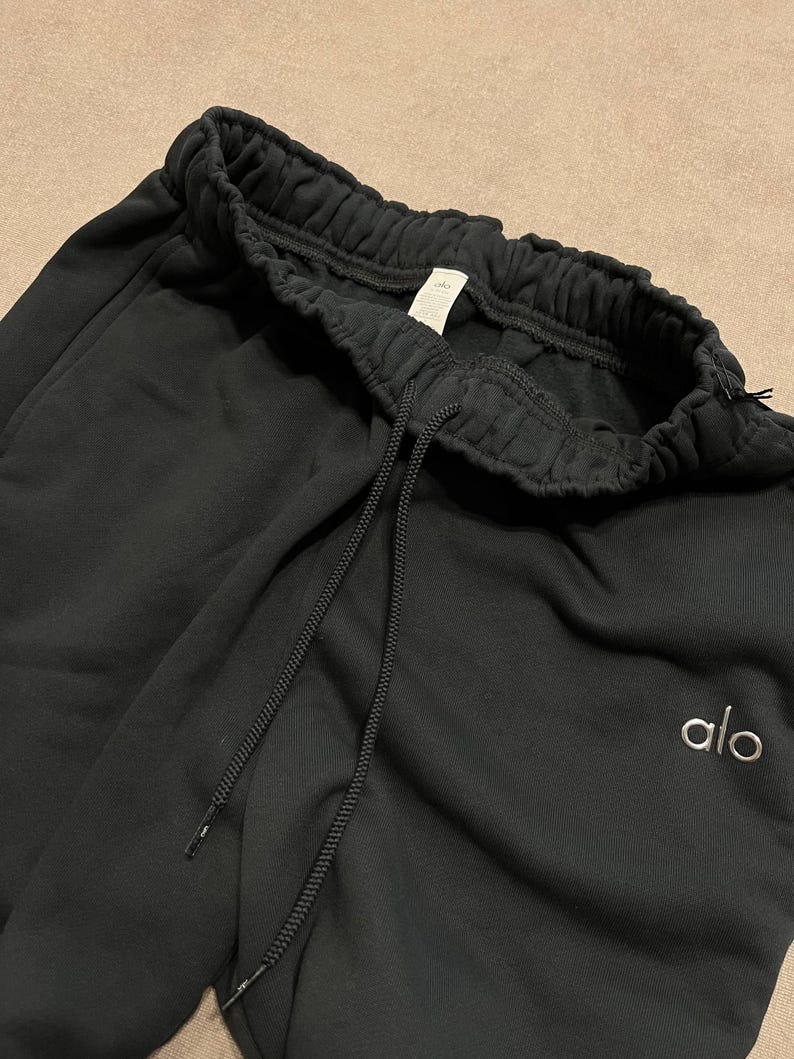 May include: Black sweatpants featuring an elasticized waistband with a matching black drawstring. The garment includes a small white embroidered logo reading alo on the left side. The fabric has a soft, matte texture and a relaxed fit design.