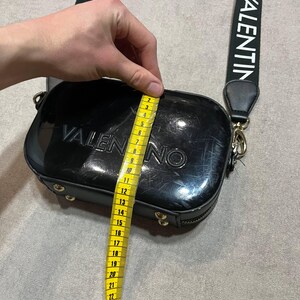 May include: Black Valentino crossbody bag with a black strap featuring the brand name in white. The bag is approximately 8 inches wide. The bag has a rectangular shape with rounded edges and gold-colored hardware.