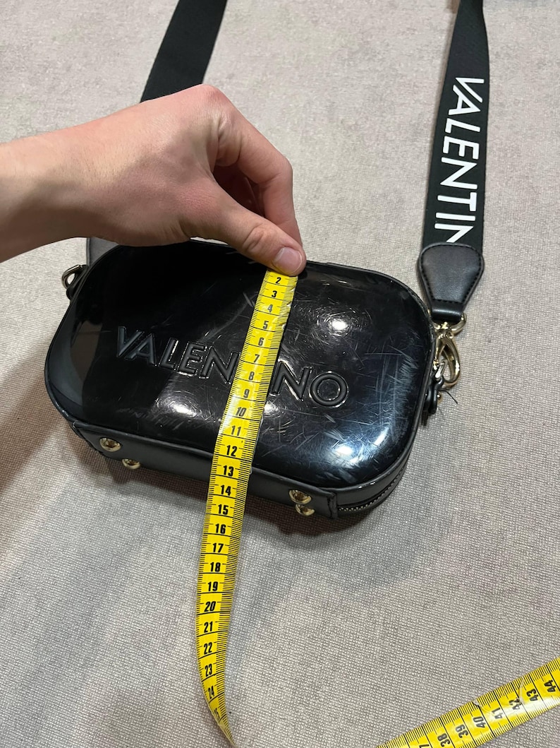 May include: Black Valentino crossbody bag with a black strap featuring the brand name in white. The bag is approximately 8 inches wide. The bag has a rectangular shape with rounded edges and gold-colored hardware.