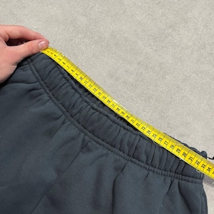 May include: Dark charcoal gray sweatpants featuring an elasticized waistband. A yellow measuring tape is positioned along the top edge of the waistband, showing a measurement of approximately 34 inches across the width of the garment.