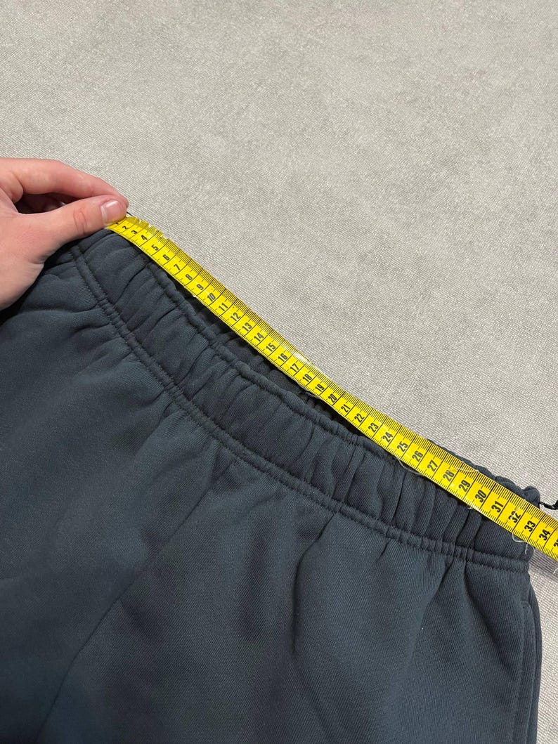 May include: Dark charcoal gray sweatpants featuring an elasticized waistband. A yellow measuring tape is positioned along the top edge of the waistband, showing a measurement of approximately 34 inches across the width of the garment.