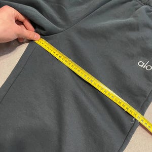 May include: Dark charcoal gray sweatpants featuring an elastic waistband and the white text logo alo. A yellow measuring tape is placed diagonally across the fabric, showing a length measurement of approximately 36 inches from the crotch seam.