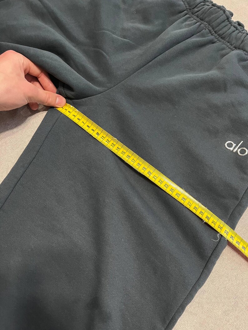 May include: Dark charcoal gray sweatpants featuring an elastic waistband and the white text logo alo. A yellow measuring tape is placed diagonally across the fabric, showing a length measurement of approximately 36 inches from the crotch seam.