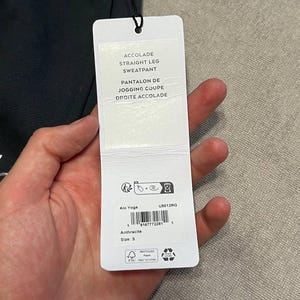 May include: A black sweatpant with a white rectangular product tag attached by a black string. The tag features the text ACCOLADE STRAIGHT LEG SWEATPANT, PANTALON DE JOGGING COUPE DROITE ACCOLADE, Alo Yoga, U5012RG, Anthracite, and size S.