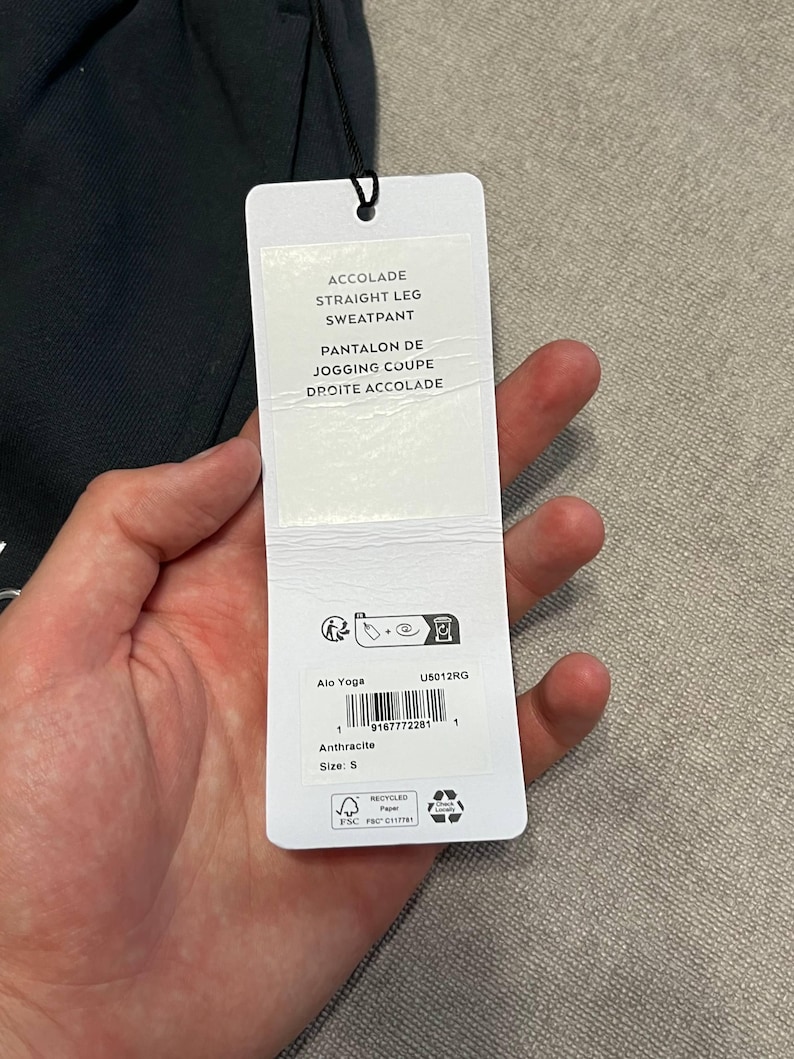 May include: A black sweatpant with a white rectangular product tag attached by a black string. The tag features the text ACCOLADE STRAIGHT LEG SWEATPANT, PANTALON DE JOGGING COUPE DROITE ACCOLADE, Alo Yoga, U5012RG, Anthracite, and size S.