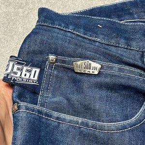 Puede incluir: Primer plano de unos vaqueros de mezclilla azul oscuro. Los vaqueros tienen costuras blancas, un bolsillo peque&ntilde;o y una etiqueta met&aacute;lica que dice "EAT 560 JOE GEAR". El bolsillo trasero tiene una etiqueta que dice "560 FIVE SIXTY".