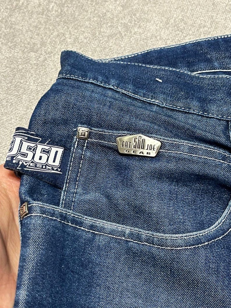 Puede incluir: Primer plano de unos vaqueros de mezclilla azul oscuro. Los vaqueros tienen costuras blancas, un bolsillo peque&ntilde;o y una etiqueta met&aacute;lica que dice "EAT 560 JOE GEAR". El bolsillo trasero tiene una etiqueta que dice "560 FIVE SIXTY".