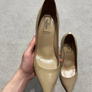 Puede incluir: Un par de zapatos de tac&oacute;n alto Valentino beige. Los zapatos tienen punta afilada y tac&oacute;n de aguja. El interior del zapato tiene una etiqueta con el logotipo de Valentino. Los zapatos son sostenidos por una mano.