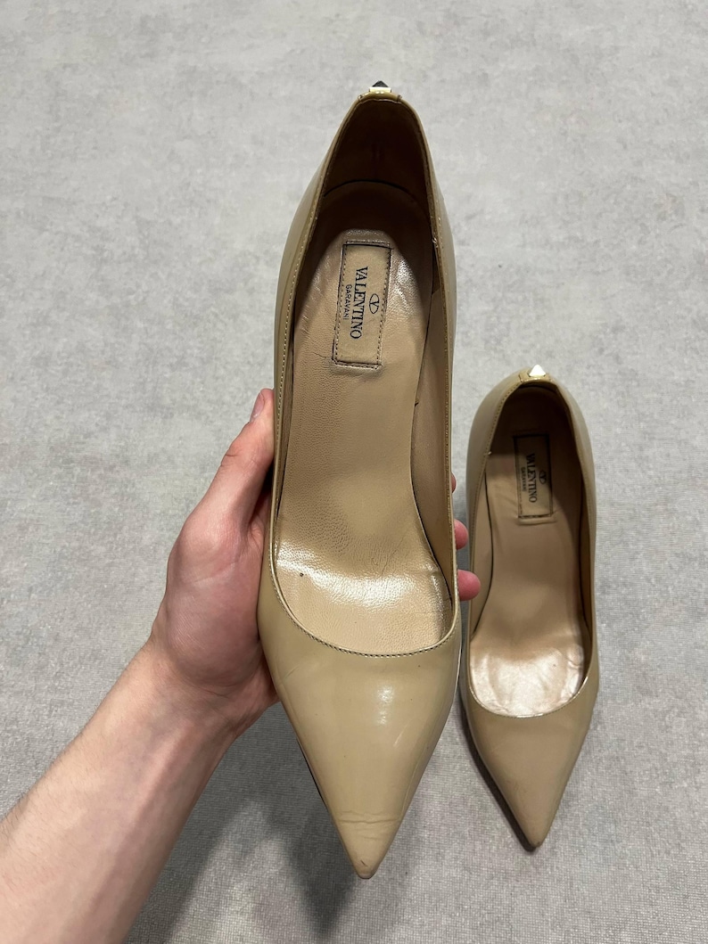 Puede incluir: Un par de zapatos de tac&oacute;n alto Valentino beige. Los zapatos tienen punta afilada y tac&oacute;n de aguja. El interior del zapato tiene una etiqueta con el logotipo de Valentino. Los zapatos son sostenidos por una mano.