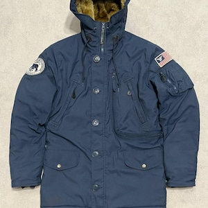 Könnte beinhalten: Marineblaue Winterparka mit fellgefütterter Kapuze. Die Jacke hat mehrere Taschen, einen Reißverschluss und Knöpfe. Auf den Ärmeln sind Aufnäher mit einem Adler und einem Bären zu sehen. Ein warmer, praktischer Mantel für kaltes Wetter.