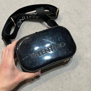 May include: Black crossbody bag with the brand name "VALENTINO" embossed on the front. The bag has a black strap with the brand name printed in white. Gold-colored hardware accents the bag.