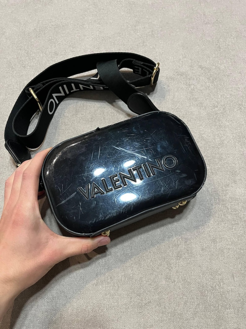 May include: Black crossbody bag with the brand name "VALENTINO" embossed on the front. The bag has a black strap with the brand name printed in white. Gold-colored hardware accents the bag.