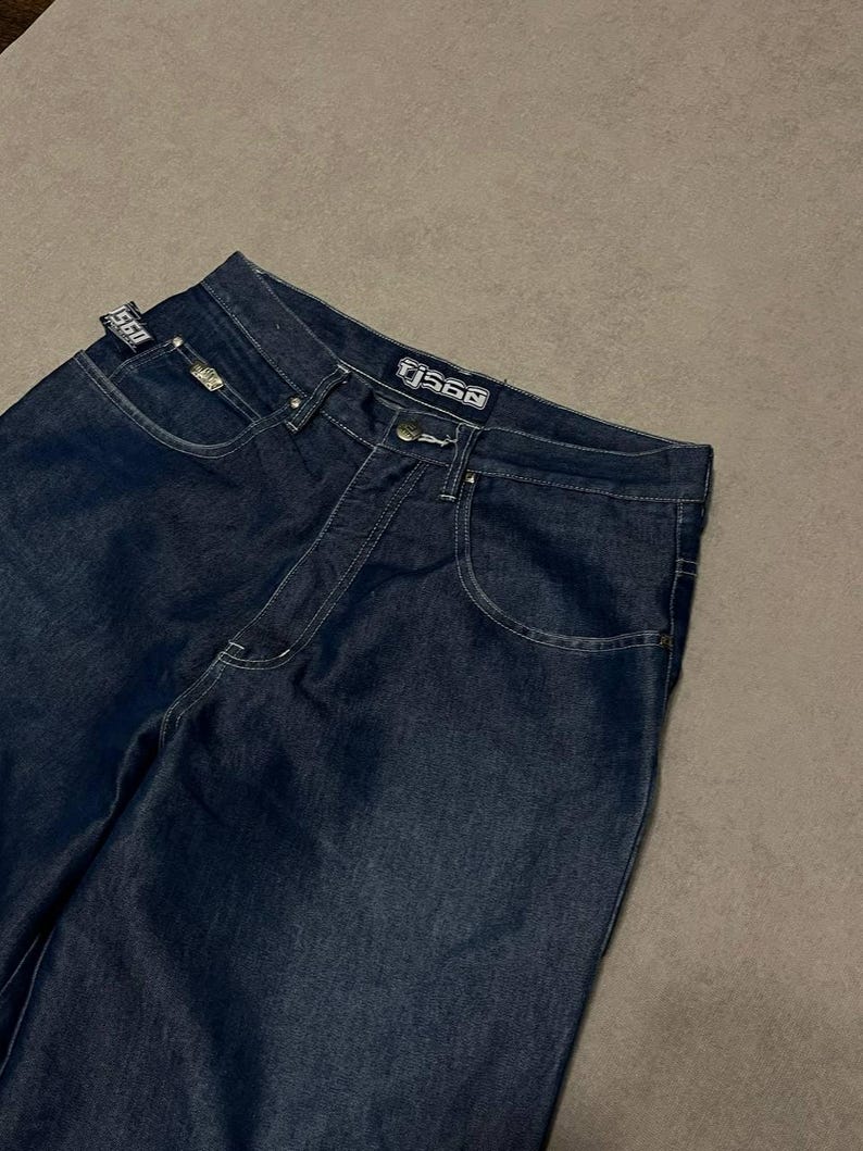 Puede incluir: Vaqueros de mezclilla azul oscuro con costuras blancas. Los vaqueros tienen un dise&ntilde;o cl&aacute;sico de cinco bolsillos y un corte de pierna recta. El nombre de la marca "TJ560" es visible en la cintura y una peque&ntilde;a etiqueta en el bolsillo delantero.