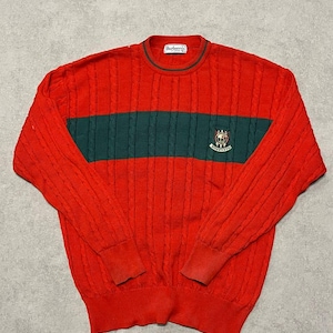 May include: Red cable-knit jumper with a green stripe across the chest. The jumper has a crew neck with green trim and a small embroidered emblem. The brand name "Burberrys" is visible on the collar.
