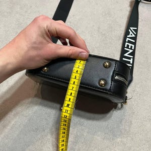 May include: Black crossbody bag with gold-colored studs and zipper. The bag's strap has the word "VALENTINO" printed in white. The bag is approximately 4 inches (10 cm) in height, as measured by a yellow tape measure.