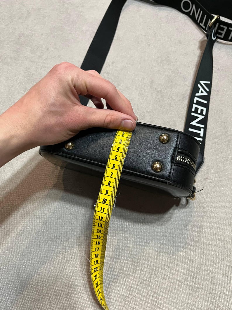 May include: Black crossbody bag with gold-colored studs and zipper. The bag's strap has the word "VALENTINO" printed in white. The bag is approximately 4 inches (10 cm) in height, as measured by a yellow tape measure.