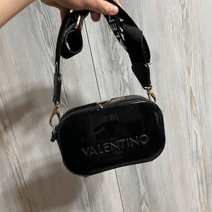 May include: A glossy black crossbody bag with the embossed word "VALENTINO" on the front. The bag has a wide black strap with white lettering and gold-colored hardware. The bag is held up by a hand.
