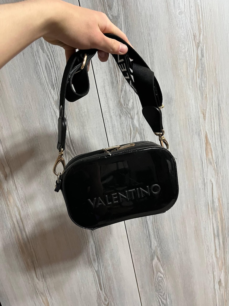 May include: A glossy black crossbody bag with the embossed word "VALENTINO" on the front. The bag has a wide black strap with white lettering and gold-colored hardware. The bag is held up by a hand.