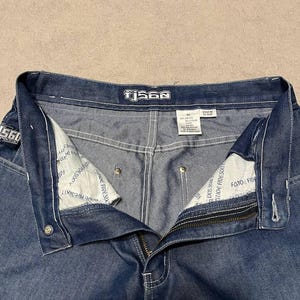 Puede incluir: Primer plano de unos vaqueros azules con cremallera y cierre de bot&oacute;n. Los vaqueros tienen costuras blancas y la marca "TjS60" es visible en la cintura. El forro interior tiene texto impreso.