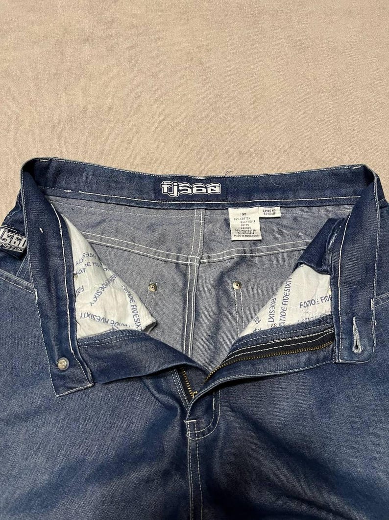 Puede incluir: Primer plano de unos vaqueros azules con cremallera y cierre de bot&oacute;n. Los vaqueros tienen costuras blancas y la marca "TjS60" es visible en la cintura. El forro interior tiene texto impreso.